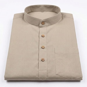 May include: A folded, beige shirt with a mandarin collar and a single pocket. The shirt has a button-down placket with three wooden buttons and a button at the collar. The shirt is made of a light-colored fabric.