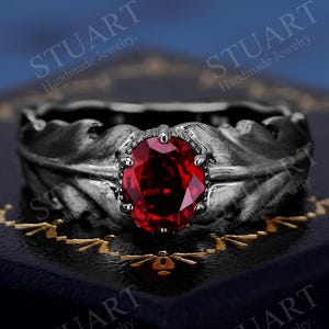May include: A silver ring with a large oval-cut ruby gemstone. The ring's band features a leaf design. The ring is displayed on a dark blue surface with gold accents. The text "STUART Handmade Jewelry" is visible.