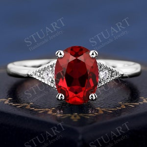 May include: A silver ring with a large oval-shaped red gemstone and two smaller diamond-shaped gemstones on either side.