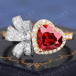 May include: A gold ring with a heart-shaped, red gemstone, surrounded by small diamonds. The ring features a bow-shaped design on one side, also encrusted with diamonds. The ring is set on a dark blue surface, with the text "STUART Handmade Jewelry".