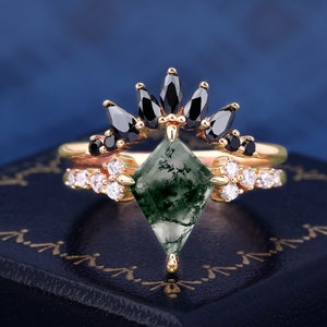 Peut inclure: Un ensemble de bagues en or avec une pierre d'agate mousse verte en forme de losange, ornée de pierres noires et blanches. L'ensemble est présenté sur une surface bleu foncé, mettant en valeur le design et le savoir-faire.