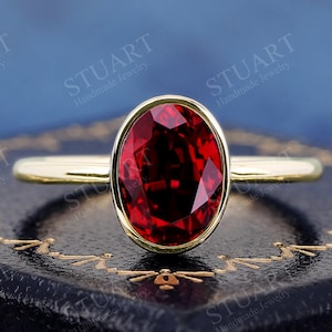 Può includere: Un anello d'oro con una grande pietra preziosa rosso scuro a taglio ovale. La pietra preziosa è incastonata in una lunetta e la fascia è in oro lucido semplice. L'anello è esposto su una superficie blu scuro.