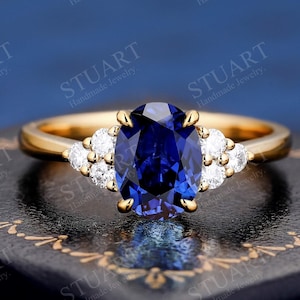 Könnte beinhalten: Ein goldener Ring mit einem großen ovalen blauen Saphir, der von kleineren weißen Diamanten umgeben ist. Der Ring liegt auf einem schwarzen Samtkasten.
