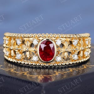 Può includere: Un anello color oro con una gemma rossa ovale al centro. L'anello presenta un design dettagliato con piccole pietre bianche e un motivo floreale. Le parole "STUART Handmade Jewelry" sono visibili.