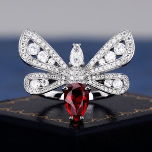 May include: A silver ring with a red gemstone in the center, shaped like a butterfly with clear stones on the wings.
