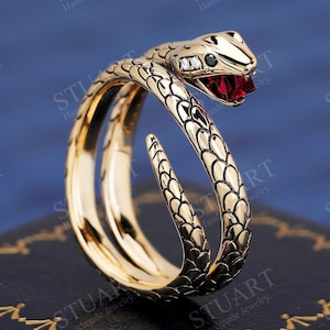 Puede incluir: Un anillo de serpiente dorado con una piedra roja para el ojo. La serpiente está enrollada alrededor de una banda y tiene un diseño texturizado y escamoso.