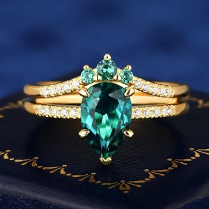 Op de afbeelding: Een verlovingsring van goud met een peervormige smaragd en een delicate band van diamanten. De smaragd is omgeven door kleinere smaragden, waardoor een kroonachtig ontwerp ontstaat.