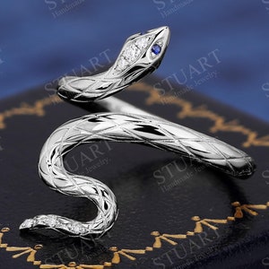 May include: A silver snake ring with a blue gemstone eye and a diamond-encrusted body. The ring is set on a black velvet background.