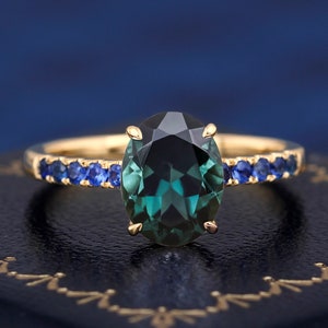 Puede incluir: Un anillo de oro con una gran piedra preciosa verde de forma ovalada rodeada por una banda de piedras azules m&aacute;s peque&ntilde;as.