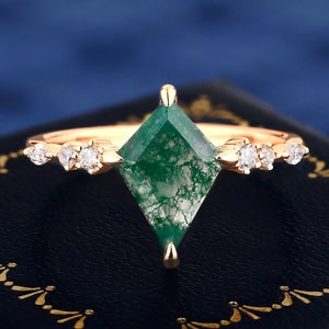 Peut inclure: Une bague en or rose ornée d'une pierre de moss agate verte en forme de losange. La bague est ornée de petites pierres précieuses rondes et claires. La bague est posée sur un fond bleu foncé.