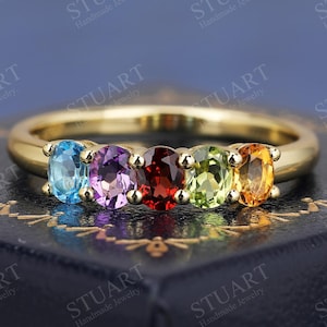 Può includere: Un anello con fascia in oro con cinque pietre preziose ovali. Le pietre preziose sono blu, viola, rosse, verdi e arancioni. L'anello è su una scatola di velluto nero.