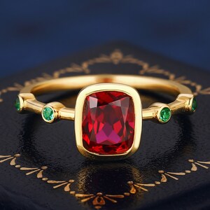 May include: A gold ring featuring a large, square-shaped red gemstone and four smaller green gemstones.