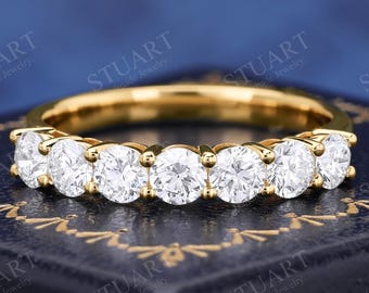 May include: A gold band ring featuring seven round, clear, faceted stones set in a row across the top. The ring is displayed on a dark blue textured box with gold decorative trim. The metal band has a polished, reflective yellow gold finish.