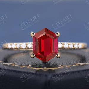 May include: A gold ring featuring a vibrant red, hexagonal gemstone as the focal point. The band is adorned with small, sparkling diamonds. The ring is set against a dark blue background.