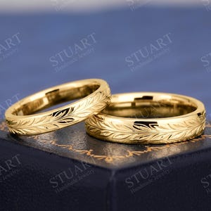 Puede incluir: Dos anillos de tono dorado con un patrón de hojas grabado de forma continua alrededor de la banda exterior. Los anillos están colocados sobre una caja azul oscuro con un borde decorativo dorado. El texto Stuart Handmade Jewelry es visible.
