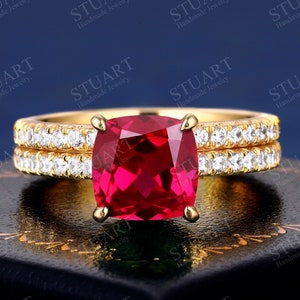 Puede incluir: Un anillo de oro con una gran piedra de rubí de talla cojín rodeada de diamantes más pequeños. El anillo está sobre una caja de terciopelo negro.