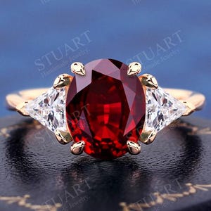 Könnte beinhalten: Ein Ring mit einem roten ovalen Edelstein und zwei kleineren dreieckigen Edelsteinen auf jeder Seite. Der Ring ist in Gold gefasst.