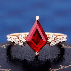 May include: A rose gold ring featuring a large, red, square-shaped gemstone and smaller, clear gemstones on the band.
