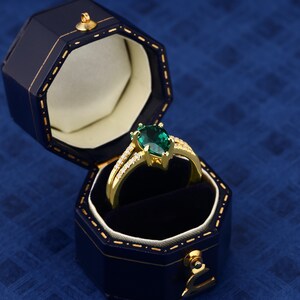 Op de afbeelding: Een gouden ring met een peervormige smaragdgroene edelsteen omringd door kleinere heldere edelstenen. De ring bevindt zich in een blauwe fluwelen ringdoos.