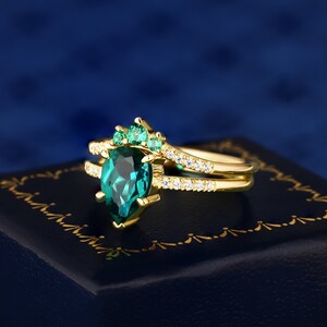 Op de afbeelding: Een gouden ring met een peervormige smaragd, omringd door kleinere smaragden. De ring heeft een delicate band met kleinere diamanten.