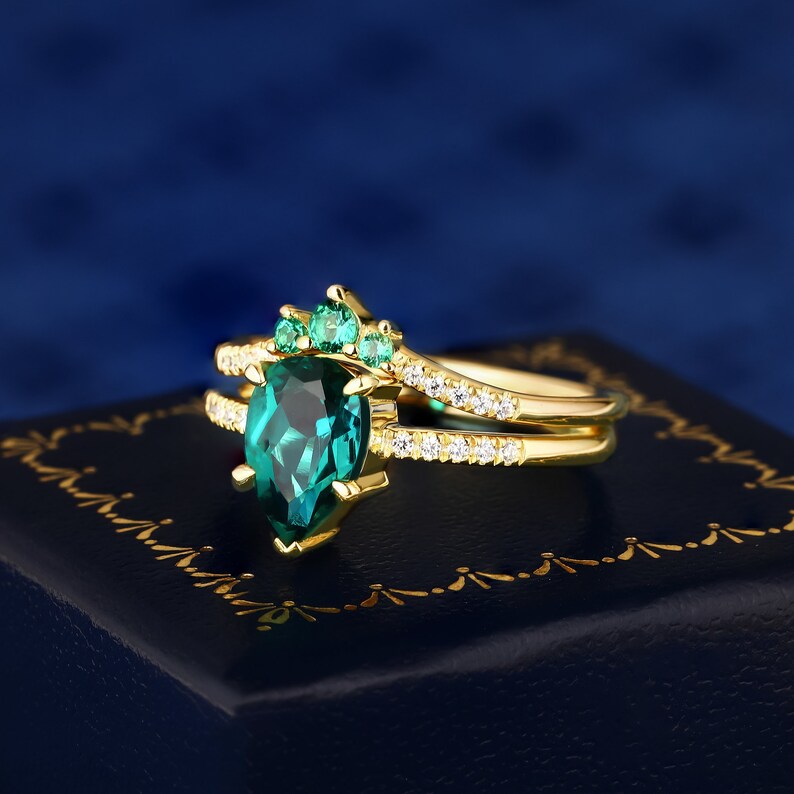 Op de afbeelding: Een gouden ring met een peervormige smaragd, omringd door kleinere smaragden. De ring heeft een delicate band met kleinere diamanten.