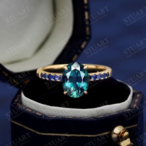 Pu&ograve; includere: Un anello d'oro con una grande pietra ovale di colore turchese circondata da una fila di piccole pietre blu. L'anello &egrave; in una scatola per anelli in velluto blu.