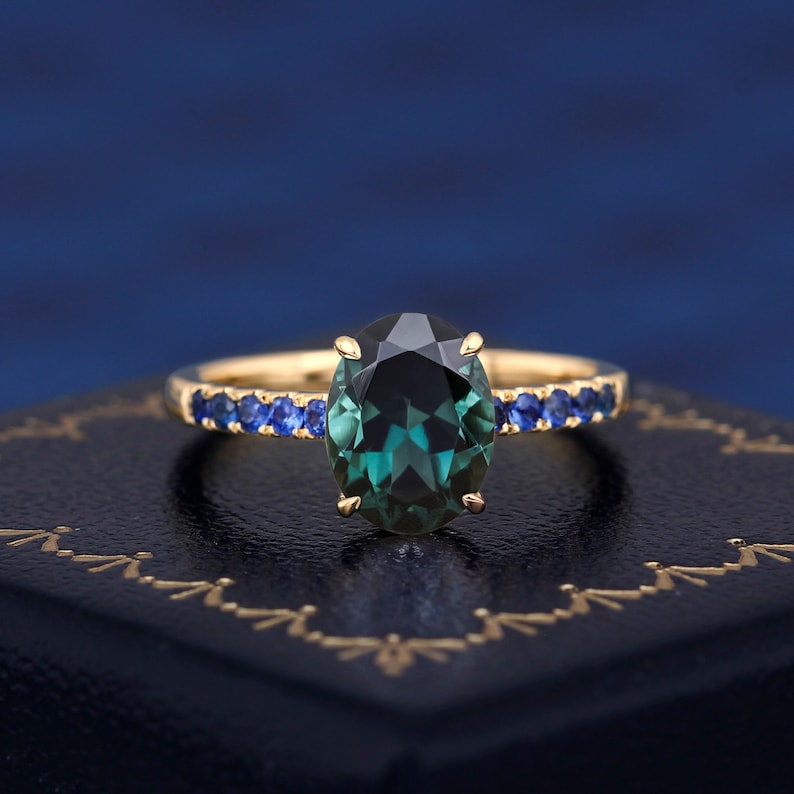 Puede incluir: Un anillo de oro con una gran piedra preciosa verde de forma ovalada rodeada por una banda de piedras azules m&aacute;s peque&ntilde;as.