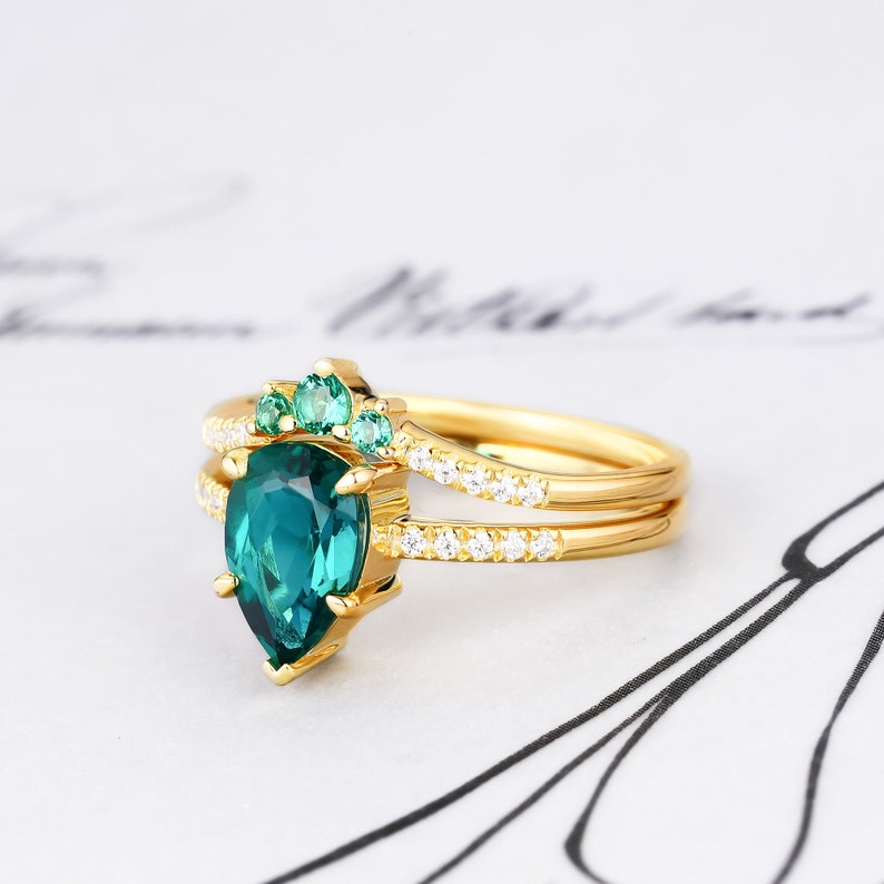 Op de afbeelding: Een gouden ring met een peervormige groene edelsteen, omringd door kleinere groene edelstenen en een band van kleine diamanten.
