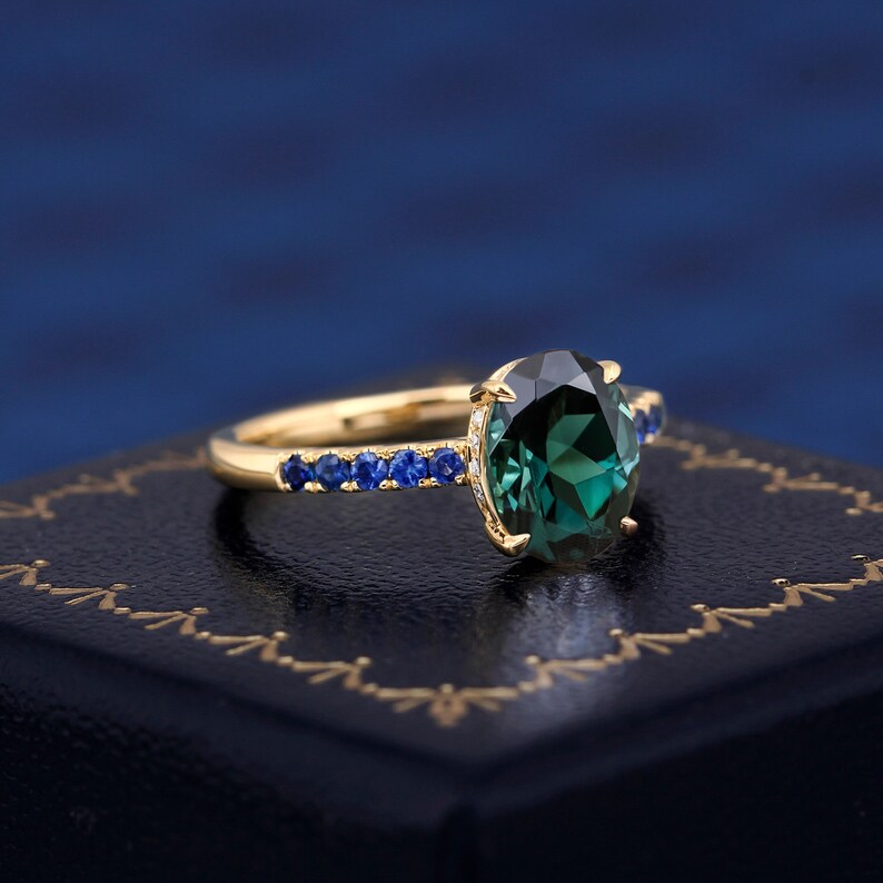 Puede incluir: Un anillo de oro con una gran piedra preciosa ovalada verde rodeada de una fila de piedras azules m&aacute;s peque&ntilde;as.