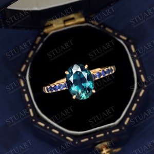 Pu&ograve; includere: Un anello d'oro con una grande pietra ovale di colore turchese circondata da una fila di pietre blu pi&ugrave; piccole. L'anello &egrave; in una scatola per anelli in velluto nero.