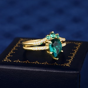 Op de afbeelding: Een gouden ring met een peervormige smaragd, omringd door kleinere smaragden. De ring heeft een delicate band met kleine diamanten.