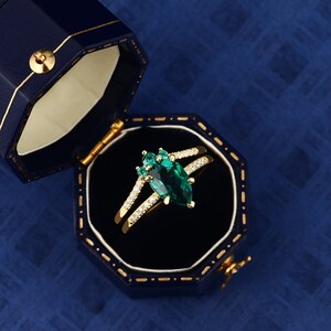 Op de afbeelding: Een gouden ring met een peervormige smaragd, omringd door kleinere smaragden en diamanten. De ring ligt in een blauwe fluwelen ringdoos.