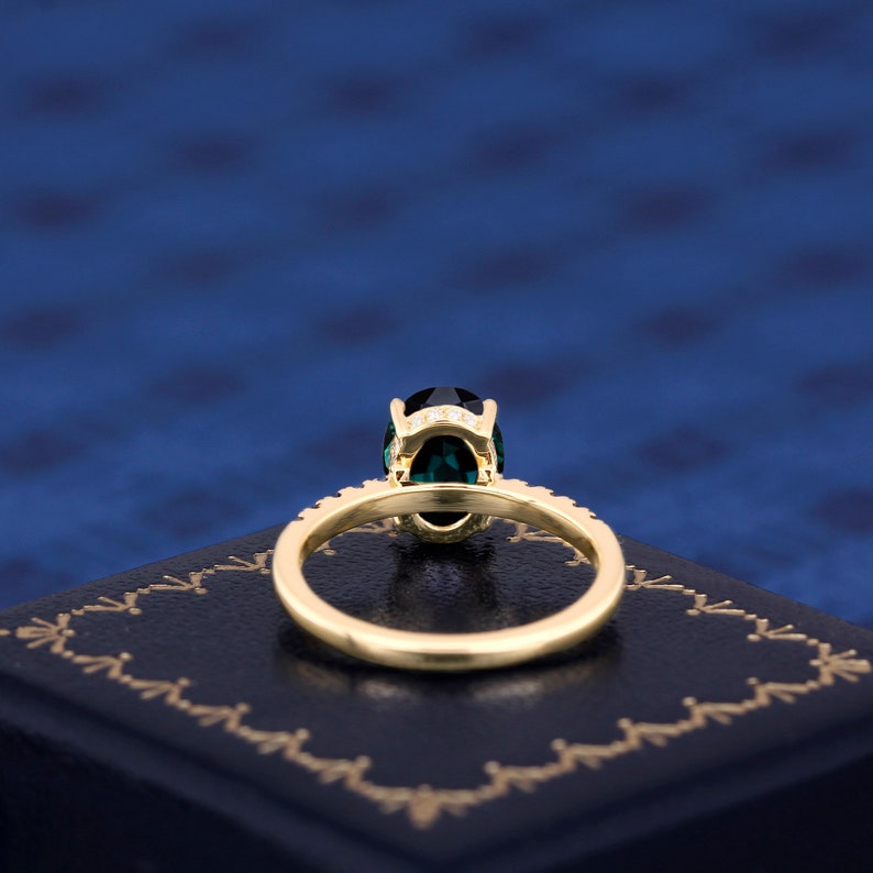 Puede incluir: Un anillo de oro con una gran piedra preciosa ovalada verde rodeada de diamantes m&aacute;s peque&ntilde;os. El anillo est&aacute; sobre una caja de terciopelo azul.