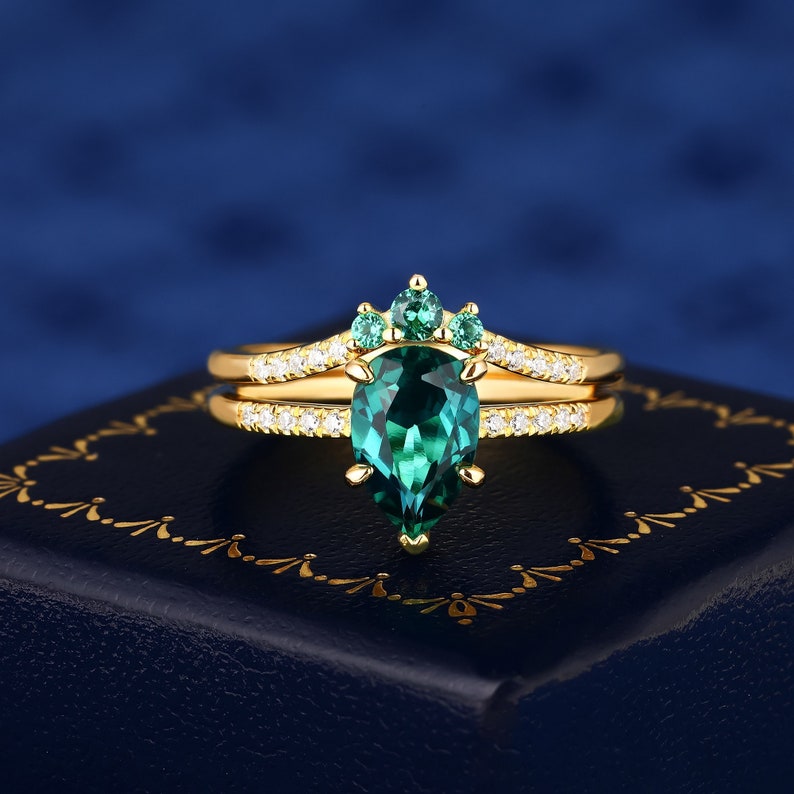 Op de afbeelding: Een verlovingsring van goud met een peervormige smaragd en een delicate band van diamanten. De smaragd is omgeven door kleinere smaragden, waardoor een kroonachtig ontwerp ontstaat.