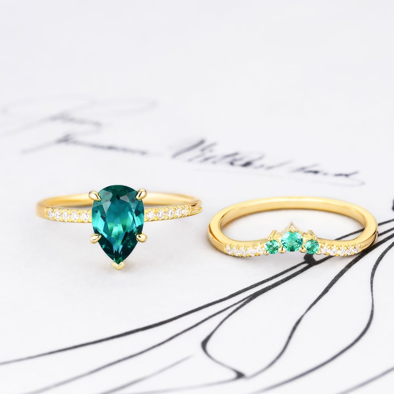 Op de afbeelding: Twee gouden ringen met smaragdgroene edelstenen en witte diamanten. De grotere ring heeft een peervormige smaragdgroene edelsteen die omringd is door witte diamanten. De kleinere ring heeft een gebogen band met drie smaragdgroene edelstenen en witte diamanten.