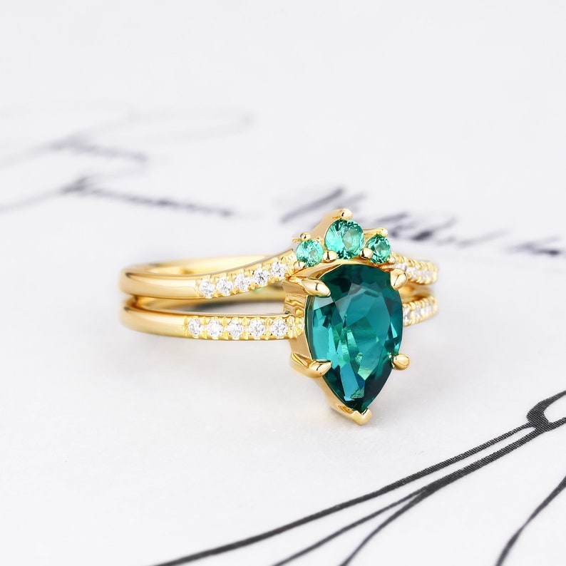 Op de afbeelding: Een gouden ring met een peervormige groene edelsteen, omringd door kleinere groene edelstenen en een band van kleine diamanten.