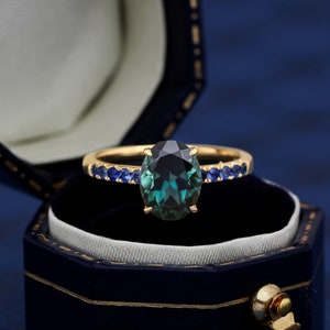 Puede incluir: Un anillo de oro con una gran piedra preciosa ovalada verde rodeada de una fila de piedras azules m&aacute;s peque&ntilde;as.