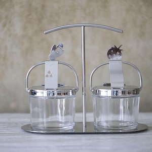 Op de afbeelding: Een roestvrijstalen kruidenset met twee heldere glazen potten. Elke pot heeft een metalen handvat en deksel. De set wordt ondersteund door een metalen standaard met een gebogen bovenkant. Eén pot is bekroond met een vlinder, de andere met een aardbei.