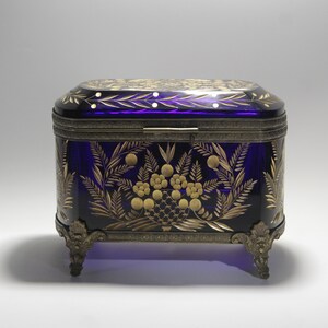Op de afbeelding: Een rechthoekige kobaltblauwe glazen doos met een scharnierend deksel, versierd met gouden bloemen- en bladerpatronen. De doos heeft een sierlijke metalen rand en rust op vier decoratieve voetjes. Het deksel en de zijkanten hebben gedetailleerde gravures.