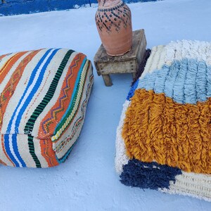 K&ouml;nnte beinhalten: Zwei farbenfrohe Bodenkissen, eines mit einem gestreiften Muster in Orange, Wei&szlig;, Blau und Gr&uuml;n, das andere mit einer flauschigen Textur in Wei&szlig;, Blau und Gelb.