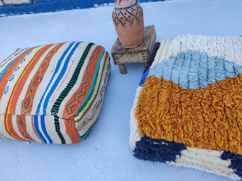 K&ouml;nnte beinhalten: Zwei farbenfrohe Bodenkissen, eines mit einem gestreiften Muster in Orange, Wei&szlig;, Blau und Gr&uuml;n, das andere mit einer flauschigen Textur in Wei&szlig;, Blau und Gelb.