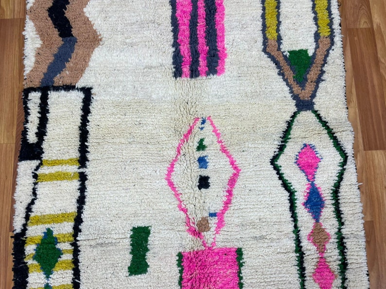 Peut inclure: Un tapis blanc avec un motif g&eacute;om&eacute;trique en noir, marron, rose, vert, jaune et bleu. Le tapis a une texture tiss&eacute;e &agrave; la main.