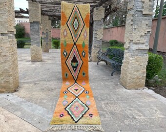 May include: A long, narrow, orange and brown rug with a geometric pattern of diamonds and triangles. The rug is laid out on a stone patio.