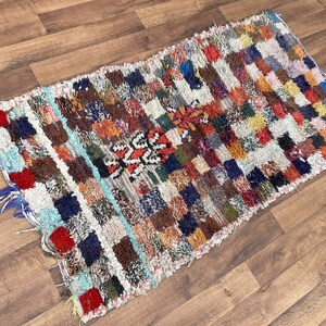 May include: A colorful, hand-woven rug with a geometric pattern in shades of red, blue, green, yellow, and brown. The rug has a frayed edge and a textured surface.