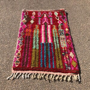 Puede incluir: Una alfombra marroqu&iacute; colorida con un patr&oacute;n geom&eacute;trico en tonos de rosa, rojo, verde, azul y marr&oacute;n. La alfombra tiene un borde de flecos y est&aacute; hecha de lana.