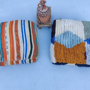 K&ouml;nnte beinhalten: Zwei farbenfrohe marokkanische Poufs, einer mit einem gestreiften Muster und der andere mit einem geometrischen Muster, sind auf einer hellblauen Oberfl&auml;che platziert. Ein kleiner Holzhocker mit einem Tontopf darauf befindet sich im Hintergrund.