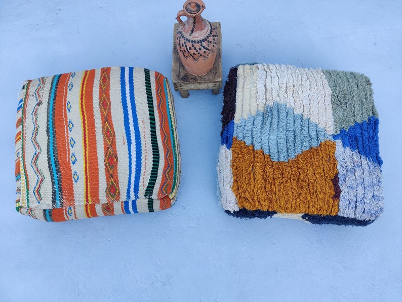 K&ouml;nnte beinhalten: Zwei farbenfrohe marokkanische Poufs, einer mit einem gestreiften Muster und der andere mit einem geometrischen Muster, sind auf einer hellblauen Oberfl&auml;che platziert. Ein kleiner Holzhocker mit einem Tontopf darauf befindet sich im Hintergrund.