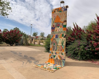 May include: A colorful, rectangular rug with geometric patterns in orange, green, blue, and gray. The rug is displayed outdoors, with a portion hanging from a metal fixture. The background includes lush greenery and a cloudy sky.