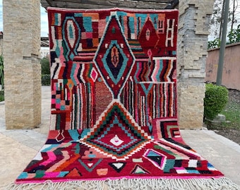 May include: A colorful Moroccan rug with a geometric pattern in shades of red, pink, blue, green, and black. The rug has a fringe border and is made of wool.