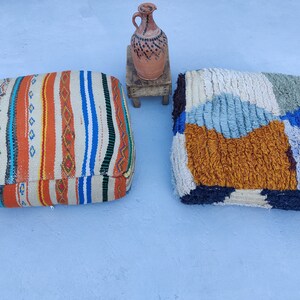 K&ouml;nnte beinhalten: Zwei bunte, gemusterte Bodenkissen, eines mit einem gestreiften Design und das andere mit einem Patchwork-Design, sind auf einer hellblauen Oberfl&auml;che abgebildet. Ein kleiner Holzhocker mit einer braunen Tonkrug darauf ist ebenfalls zu sehen.